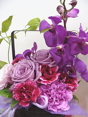 紫系のバラやカーネーション、蘭、トルコキキョウを竹の取っ手付きバスケットにまとめたアレンジメント